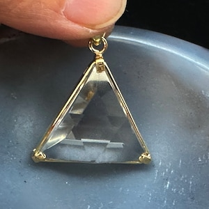 May include: A gold-toned pendant featuring a clear triangular crystal with a gold-toned frame. The pendant has a loop at the top for attaching to a chain.