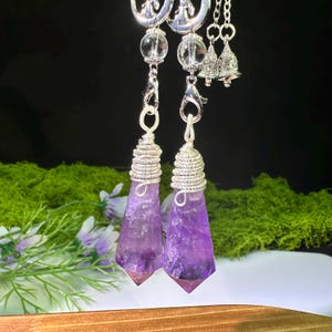 May include: Two amethyst crystal pendants wrapped in silver-toned wire. Each pendant features a clear bead, a silver crescent moon charm, and a small silver bell attached to a metal chain. The purple crystals are pointed at the bottom.