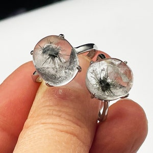 K&ouml;nnte beinhalten: Zwei Ringe mit klaren Quarzsteinen mit schwarzen Einschl&uuml;ssen. Die Ringe haben silberne B&auml;nder. Die klaren Quarzsteine sind rund und haben ein stachelartiges Sternmuster. Die Ringe werden von einer Hand gehalten.