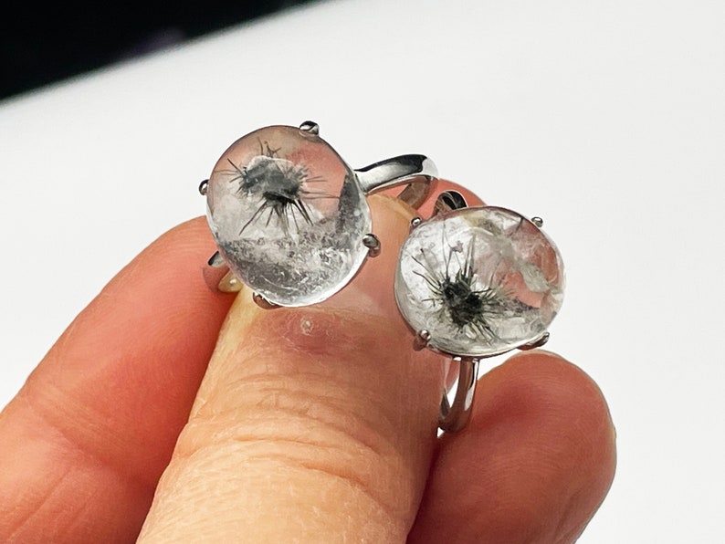K&ouml;nnte beinhalten: Zwei Ringe mit klaren Quarzsteinen mit schwarzen Einschl&uuml;ssen. Die Ringe haben silberne B&auml;nder. Die klaren Quarzsteine sind rund und haben ein stachelartiges Sternmuster. Die Ringe werden von einer Hand gehalten.