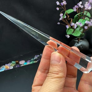 Könnte beinhalten: Ein klarer Quarz-Kristallstab mit einer Spitze und einem abgerundeten Boden. Der Stab wird in einer Hand vor einem dunklen Hintergrund gehalten, mit einer kleinen dekorativen Pflanze im Hintergrund.