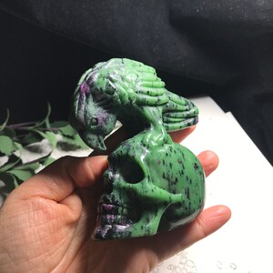 May include: A green and black gemstone skull sculpture with a bird perched on top. The skull is carved with intricate details and the bird has a detailed wingspan.