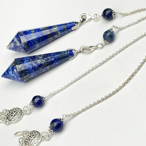May include: Two lapis lazuli crystal pendulums with silver chains and beads. The pendulums are pointed and have a dark blue color with gold flecks.