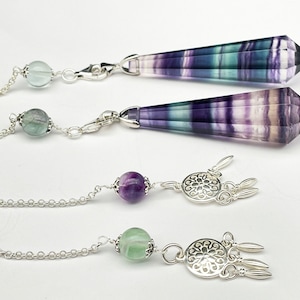 May include: Two silver chain pendulums with fluorite crystal points. The top pendulum has a green fluorite bead, the bottom pendulum has a purple fluorite bead. Both pendulums have silver dreamcatcher charms.