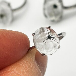K&ouml;nnte beinhalten: Ein klarer Quarzring mit schwarzen Einschl&uuml;ssen. Der ovale Edelstein ist in einem silberfarbenen Band gefasst. Der Ring wird von den Fingern einer Person gehalten, wobei weitere Ringe im Hintergrund sichtbar sind. Der Ring ist ein Schmuckst&uuml;ck.