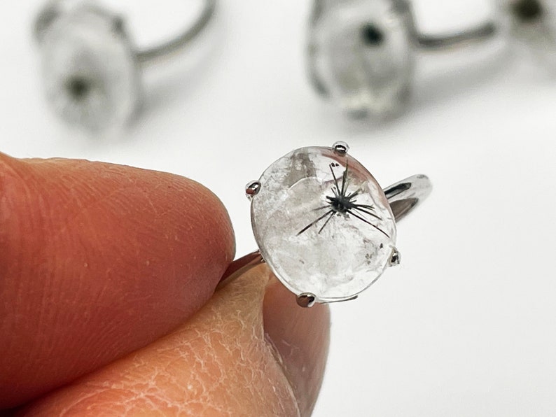 K&ouml;nnte beinhalten: Ein klarer Quarzring mit schwarzen Einschl&uuml;ssen. Der ovale Edelstein ist in einem silberfarbenen Band gefasst. Der Ring wird von den Fingern einer Person gehalten, wobei weitere Ringe im Hintergrund sichtbar sind. Der Ring ist ein Schmuckst&uuml;ck.
