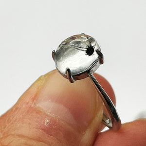 K&ouml;nnte beinhalten: Ein silberner Ring mit einem klaren, ovalen Edelstein mit einem schwarzen Einschluss. Der Edelstein ist in einer Vier-Zacken-Fassung gefasst, und der Ring hat einen schlanken, polierten Schaft. Wei&szlig;er Hintergrund.