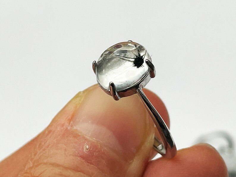 K&ouml;nnte beinhalten: Ein silberner Ring mit einem klaren, ovalen Edelstein mit einem schwarzen Einschluss. Der Edelstein ist in einer Vier-Zacken-Fassung gefasst, und der Ring hat einen schlanken, polierten Schaft. Wei&szlig;er Hintergrund.