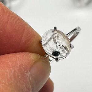 K&ouml;nnte beinhalten: Silberring mit einem ovalen, transparenten Stein, der schwarze und wei&szlig;e Einschl&uuml;sse aufweist, die an einen L&ouml;wenzahnsamenstand erinnern. Der Ring hat ein schlichtes Design mit einem schmalen Band und vier Krappen.