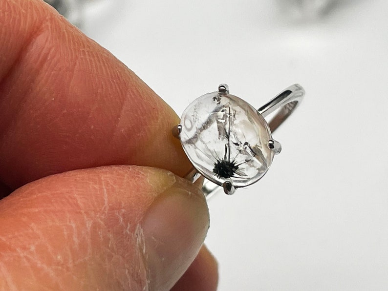 K&ouml;nnte beinhalten: Silberring mit einem ovalen, transparenten Stein, der schwarze und wei&szlig;e Einschl&uuml;sse aufweist, die an einen L&ouml;wenzahnsamenstand erinnern. Der Ring hat ein schlichtes Design mit einem schmalen Band und vier Krappen.