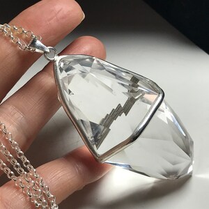 May include: A clear quartz crystal pendant with a silver setting, hanging from a silver chain. The crystal is faceted and has a pointed tip.