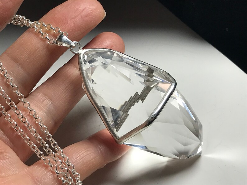 May include: A clear quartz crystal pendant with a silver setting, hanging from a silver chain. The crystal is faceted and has a pointed tip.