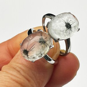 K&ouml;nnte beinhalten: Zwei silberne Ringe, jeweils mit einem klaren, ovalen Stein mit einem schwarzen, stacheligen Einschluss. Die Ringe werden vor einem wei&szlig;en Hintergrund pr&auml;sentiert, um das einzigartige Design und die Handwerkskunst des Schmucks hervorzuheben.