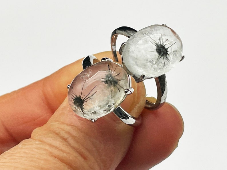 K&ouml;nnte beinhalten: Zwei silberne Ringe, jeweils mit einem klaren, ovalen Stein mit einem schwarzen, stacheligen Einschluss. Die Ringe werden vor einem wei&szlig;en Hintergrund pr&auml;sentiert, um das einzigartige Design und die Handwerkskunst des Schmucks hervorzuheben.