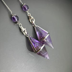 Peut inclure: Deux chaînes en métal argenté ornées de perles rondes en améthyste violette et de pendentifs en cristal d'améthyste facetté en forme de pointe, enchâssés dans des cadres en métal argenté. Les pendentifs sont fixés par des fermoirs mousquetons.