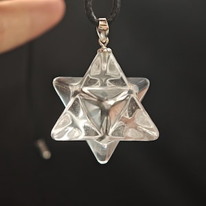 May include: Clear crystal Merkaba pendant with a silver-coloured bail and a black cord. The pendant is a three-dimensional star shape with triangular facets. The background is a solid black.