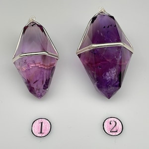 Könnte beinhalten: Zwei Amethyst-Kristall-Anhänger, einer größer als der andere, sind in Silberdraht gefasst. Die Kristalle sind tiefviolett und haben eine spitze Form. Die Anhänger sind mit den Nummern 1 und 2 gekennzeichnet.