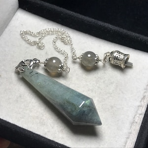 May include: A silver-colored pendulum with a faceted, blue-green crystal point. The pendulum has a chain with two gray gemstone beads and a small bell-shaped charm. The crystal point is approximately 3 inches long.