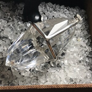 May include: A clear quartz crystal pendant encased in a silver cage, resting on a bed of white quartz stones.