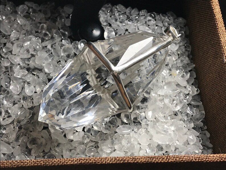 May include: A clear quartz crystal pendant encased in a silver cage, resting on a bed of white quartz stones.