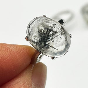 K&ouml;nnte beinhalten: Ein silberner Ring mit einem klaren, ovalen Edelstein mit schwarzen Einschl&uuml;ssen. Der Ring hat ein silbernes Band und vier Krappen. Der Ring wird von einem Finger gehalten.
