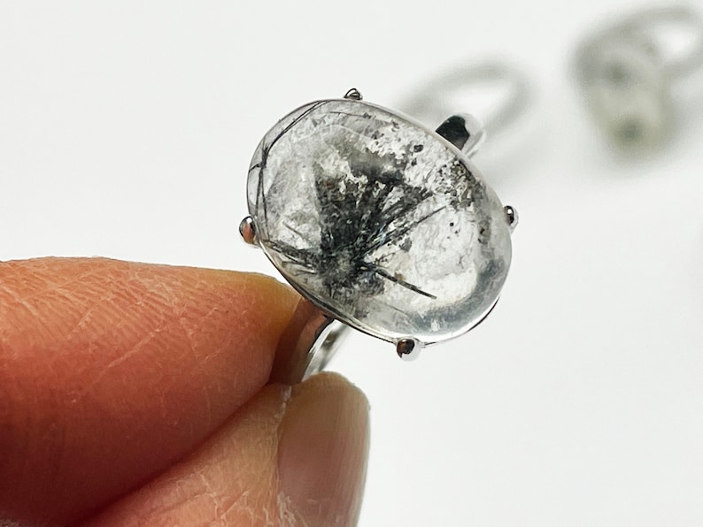 K&ouml;nnte beinhalten: Ein silberner Ring mit einem klaren, ovalen Edelstein mit schwarzen Einschl&uuml;ssen. Der Ring hat ein silbernes Band und vier Krappen. Der Ring wird von einem Finger gehalten.