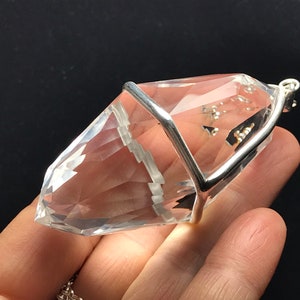 May include: A clear quartz crystal pendant with a silver cage setting. The crystal is faceted and has a pointed tip. The pendant is attached to a silver chain.