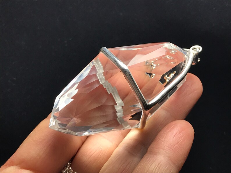 May include: A clear quartz crystal pendant with a silver cage setting. The crystal is faceted and has a pointed tip. The pendant is attached to a silver chain.