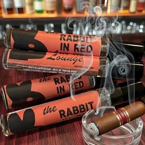 May include: Image of several glass tubes with red labels and black rabbit silhouettes, stacked near a lit cigar in a glass ashtray. The labels read "RABBIT IN RED Lounge" and "the RABBIT IN RED". Smoke rises from the cigar.