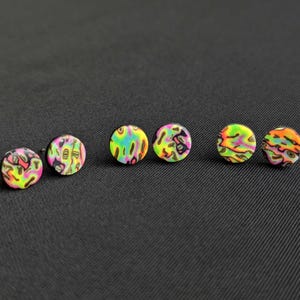 May include: Five colourful round earrings with a black background. The earrings have a psychedelic pattern with pink, green, yellow, and black colours.