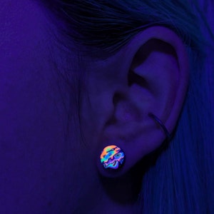 May include: A close-up of a person's ear with a colourful, iridescent ear gauge. The gauge is a round, flat disc with a swirling pattern of pink, blue, green, and black.