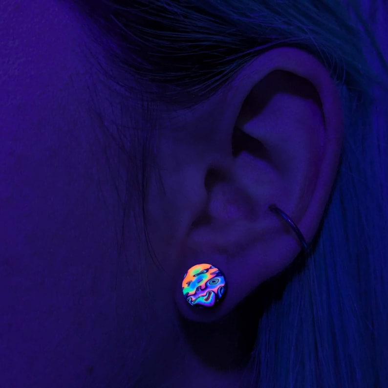 May include: A close-up of a person's ear with a colourful, iridescent ear gauge. The gauge is a round, flat disc with a swirling pattern of pink, blue, green, and black.