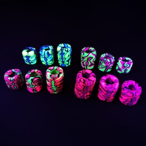 Può includere: Una collezione di 10 perline cilindriche colorate e con motivi. Le perline sono realizzate in un materiale liscio e lucido e presentano una varietà di colori vivaci, tra cui rosa, verde e blu. Le perline sono disposte a semicerchio su uno sfondo nero.