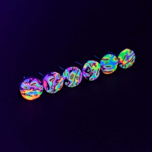 May include: Six iridescent, round, black-rimmed, glow-in-the-dark earrings with a swirling, abstract pattern in shades of pink, blue, green, and yellow.