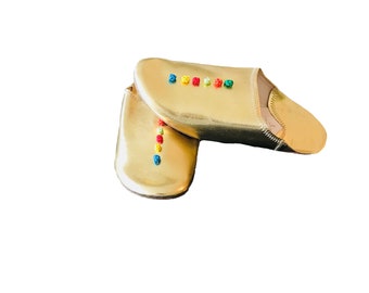 Puede incluir: Un par de zapatillas de cuero doradas con detalles bordados de colores. Las zapatillas son de punta y tienen suela plana.