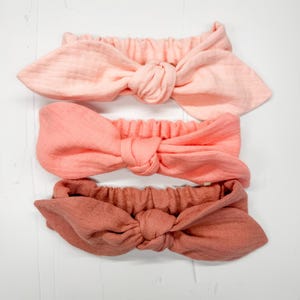 May include: Three pastel-coloured headbands with knotted bows. The headbands are in shades of light pink, coral, and terracotta. The fabric appears to be a soft, textured material. The headbands are arranged on a white surface.