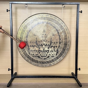 May include: A large circular bronze gong suspended from a black metal frame. The gong features intricate gold-colored engravings of a multi-headed deity and decorative patterns. A mallet with a red felt head is positioned to strike the gong surface.