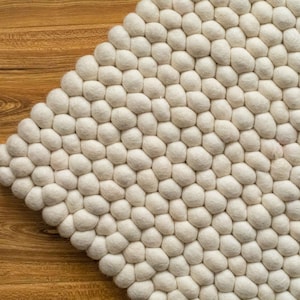 Puede incluir: Una alfombra redonda y esponjosa de color blanco, hecha de bolas de lana. La alfombra está hecha de muchas pequeñas bolas redondas de lana que están cosidas juntas para crear una superficie suave y texturizada.
