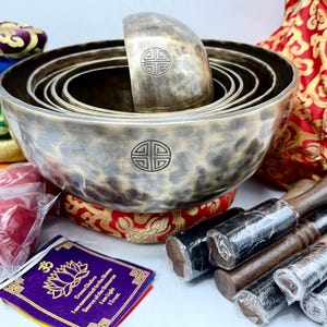 Puede incluir: Un conjunto de siete cuencos cantores con un acabado martillado, anidados uno dentro del otro. Los cuencos son de metal y tienen un color marrón oscuro. El conjunto incluye dos mazos de madera con puntas de goma negra y una pequeña bolsa de tela morada con un diseño de flor de loto y texto en un idioma extranjero. El texto dice "Cuencos cantores tibetanos genuinos, Armonía del universo, Sonido del corazón, 17,5 cm de alto".
