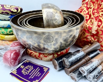May include: A set of seven singing bowls with a hammered finish, nested inside each other. The bowls are made of metal and have a dark brown color. The set includes two wooden mallets with black rubber tips and a small purple fabric pouch with a lotus flower design and text in a foreign language. The text reads "Genuine Tibetan Singing Bowls, Harmony of the Universe, Sound of the Heart, 17.5cm High".