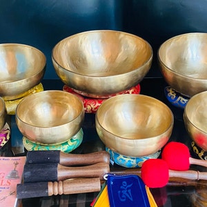 Puede incluir: Un conjunto de cuencos tibetanos de latón de varios tamaños, colocados sobre cojines coloridos. Los cuencos se acompañan de mazos de madera con cabezas de fieltro rojo, un libro titulado "Singing Bowls" y una bandera de oración.