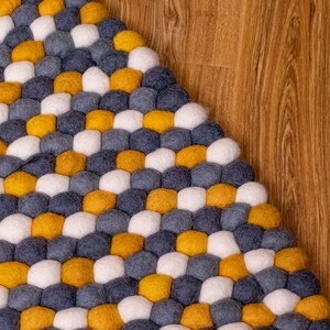 May include: A round, multi-colored rug made of felt balls. The rug is made up of yellow, white, and gray felt balls arranged in a pattern.