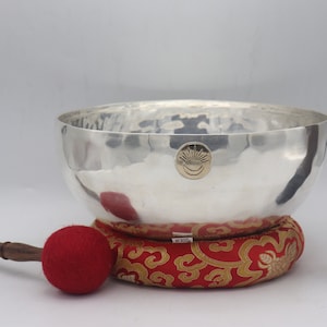 May include: A silver singing bowl with a hammered finish, a red and gold cushion, a wooden mallet with a black head, and a wooden mallet with a red head.