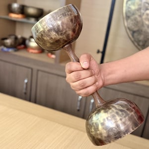 Puede incluir: Un instrumento con forma de mancuerna que presenta dos cuencos metálicos de tono bronce conectados por un mango central de madera. Cada cuenco tiene un acabado liso y desgastado, y muestra un pequeño emblema circular grabado con un patrón de cruz geométrica.