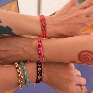 Puede incluir: Tres pulseras tejidas con texto personalizado. La pulsera superior es de color coral con la palabra FILOU. La pulsera central es rosa con la palabra MAMA. La pulsera inferior es de color marrón oscuro con la palabra MAMMA, acompañada de una cadena dorada y una pulsera de cuentas azules.