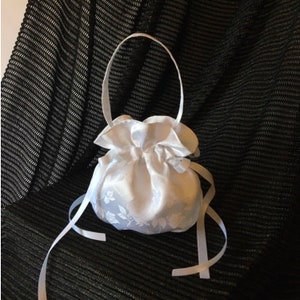 May include: A white satin drawstring bag with a handle and ribbon ties. The bag has a floral pattern and is gathered at the top. The background is a black textured fabric.