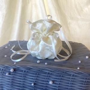 May include: Ivory satin drawstring pouch with a floral pattern. The pouch is tied with white ribbons and sits on a gray surface with scattered pearls. The background features a cream-colored curtain.