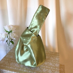 May include: A light green satin knot bag with a single handle. The bag is made of a soft, silky fabric and has a simple, elegant design.