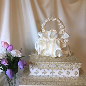 May include: A white satin drawstring bag with a ruffled handle. The bag is sitting on a stack of boxes with a floral pattern.