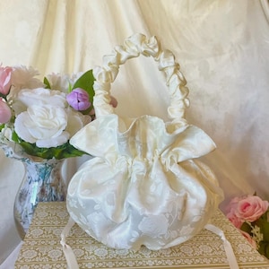 May include: Ivory satin drawstring bag with a floral pattern. The bag has a gathered handle and a ruffled top. A bouquet of flowers is in the background.